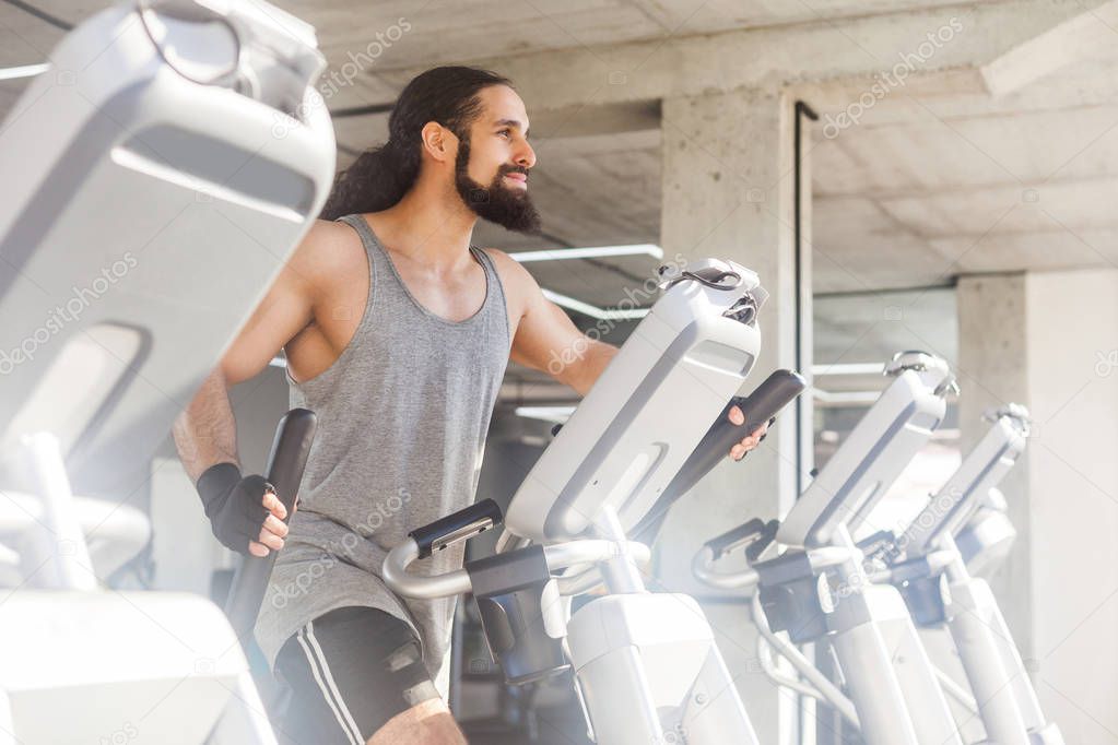 side view of young adult sportsman with long curly hair training on elliptical machine in gym, fitness concept