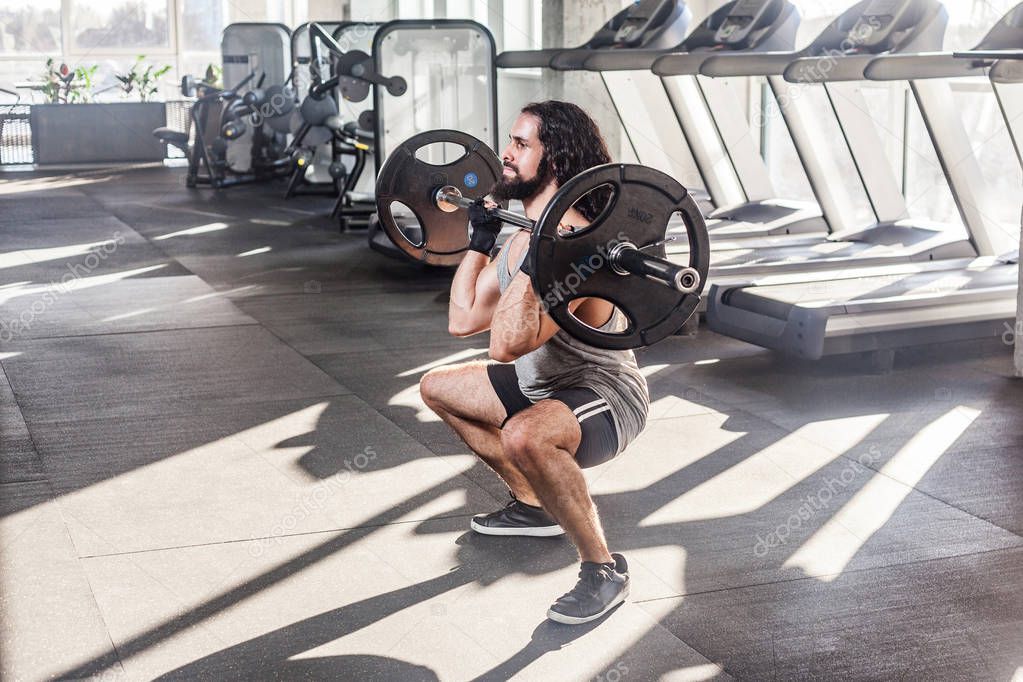 Side view of young adult bodybuilder with long curly hair working out at gym while lifting barbell and doing exercises for legs, healthy lifestyle concept  