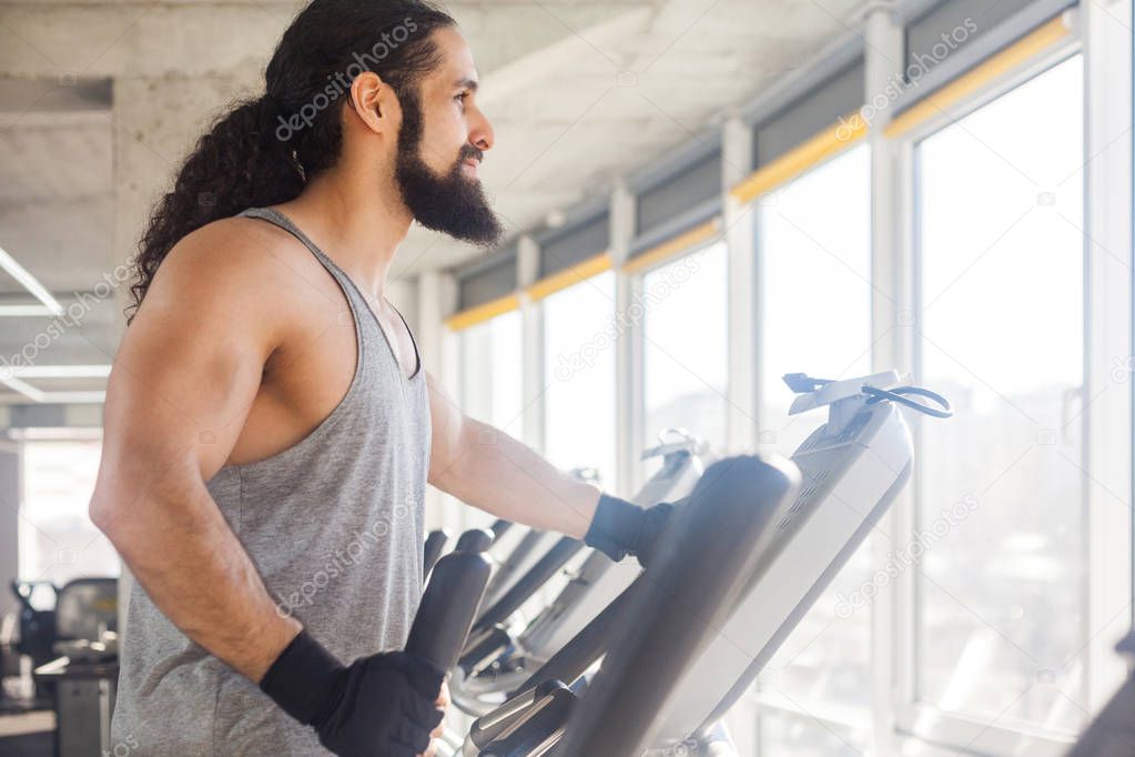 side view of young adult sportsman with long curly hair training on elliptical machine and looking at window in gym, fitness concept