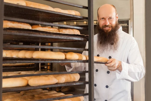 smiling adult baker with long beard in white uniform standing at workplace and pushing baking tray of bread in kitchen of  bakery, profession concept