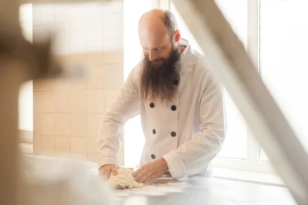Adult baker with long beard in white uniform standing at workplace and prepare bread dough with hands kneading dough on table with flour powder, concept of baking