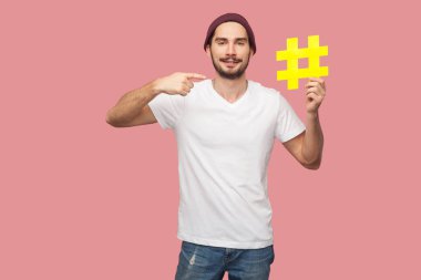Beyaz t-shirt ve rahat şapka sarı hashtag tutan ve pembe arka plan üzerinde parmak kendini işaret mutlu yakışıklı sakallı genç hipster adam Portresi 