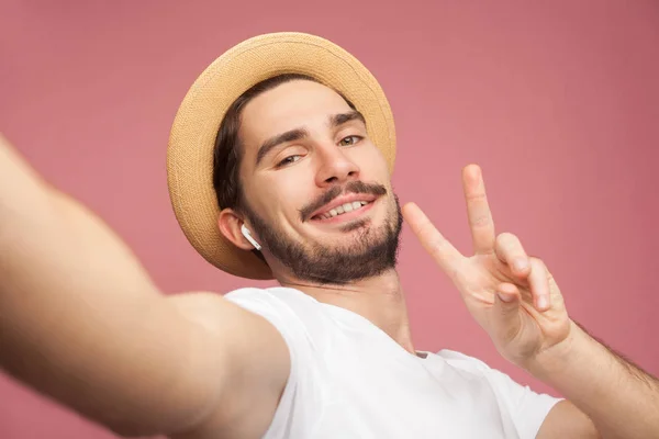 白いTシャツとカジュアルな帽子のポーズとピンクの背景にピースサインを示しながら 携帯電話で自分撮りを作るファッションハンサムなひげの若いヒップスターブロガー — ストック写真