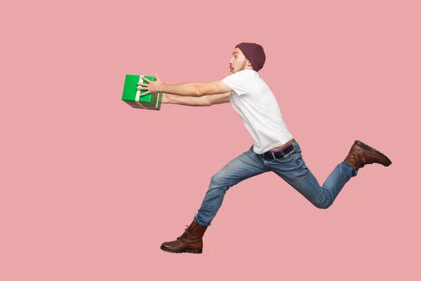 Side view of crazy bearded young hipster man in white  t-shirt and casual hat running and hurry up with delivery green present box on pink background