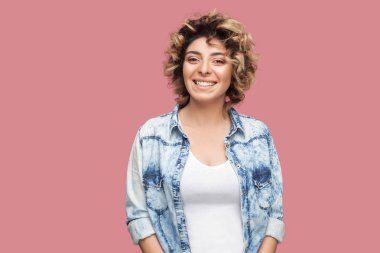 Makyaj ve rahat mavi gömlek ayakta kıvırcık saç modeli ile güzel mutlu genç kadın Portresi ayakta ve pembe arka plan üzerinde dişli gülümseme ile kamera bakıyor
