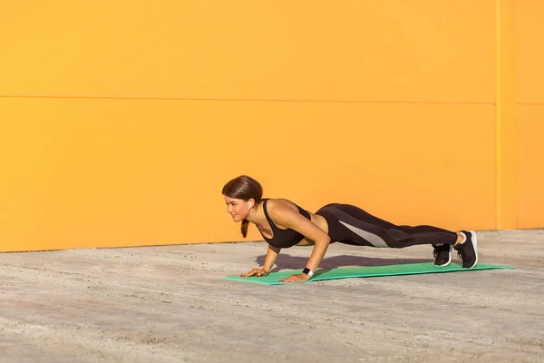 オレンジ色の壁背景に照らされた緑のマットの板の位置に立つ黒いスポーツウェアの筋肉スポーティな強い若い運動女性の側面図 チャトゥランガダンダサナヨガの姿勢 — ストック写真