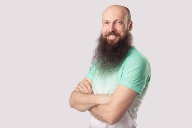 açık yeşil t-shirt uzun sakallı mutlu memnun orta yaşlı kel adam çapraz kolları ile ayakta ve gri arka plan üzerinde dişli gülümseme ile kamera bakıyor