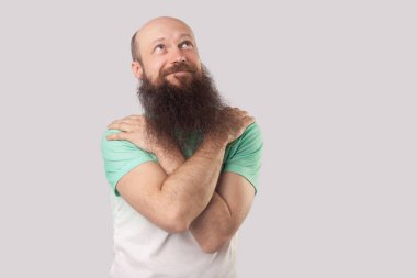 açık yeşil t-shirt uzun sakallı rüya orta yaşlı kel adam kendini sarılma ve gri arka plan üzerinde rüya görürken uzağa bakıyor