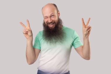 gri arka planda kamera bakarken ve göz kırpırken zafer jesti gösteren açık yeşil t-shirt uzun sakallı komik orta yaşlı kel adam