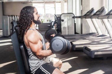 Uzun kıvırcık saçlı genç yetişkin adamın yan görünümü spor salonunda çalışırken bankta otururken ve kolları kaldırıp iki halter tutarken, pazı için egzersizler yapıyor 
