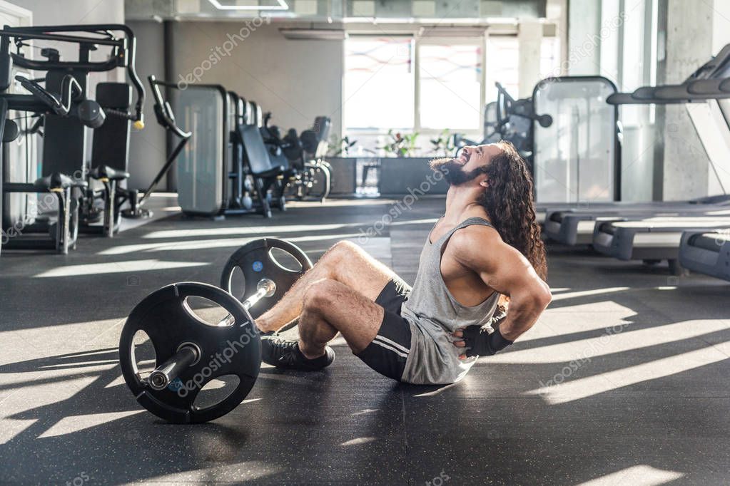 injury young adult man athlete with long curly hair working out in gym while sitting on floor and having strong hurt problem with back, spasm painful 