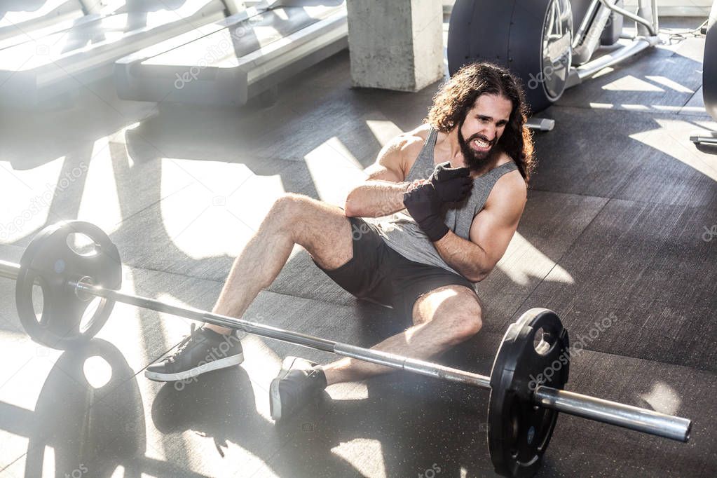 Portrait of screaming young adult man athlete with long curly hair working out in gym and screaming while sitting on floor and have strong hurt problem with wrist, spasm painful 