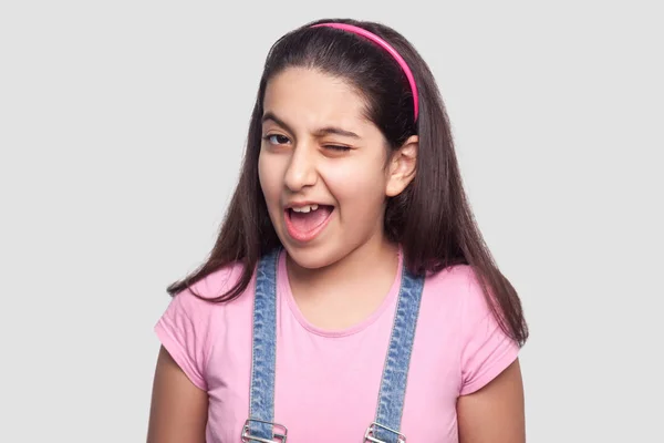 Funny beautiful brunette young girl in casual pink t-shirt and blue denim overalls winking and looking at camera on light gray background — Stock Image Funny Beautiful Brunette Young Girl Casual Pink Shirt Blue Denim Royalty Free Stock Photos