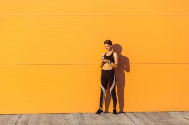 güneş ışığı turuncu duvar arka plan yakınında dururken akıllı telefon kullanarak siyah spor giyim genç mutlu çekici sportif kadın 