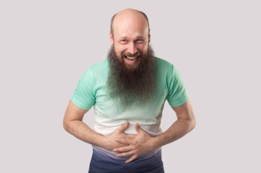 açık yeşil t-shirt uzun sakallı komik orta yaşlı kel adam karnını tutarak ve gri arka planda kameraya bakarken gülüyor