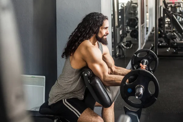 Side view of young adult bodybuilder with long curly hair preparing to lifting barbell while doing exercises for biceps in gym