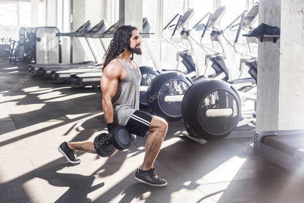 Side view of concentration young adult sportman athlete with long curly hair squatting on knee with dumbbell while doing exercises for legs in gym