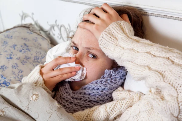若いです白いセーターと青いスカーフで彼女の鼻をきれいにし 痛みを伴う頭を保持しながら寒さを感じます病気とベッドの中で家 — ストック写真