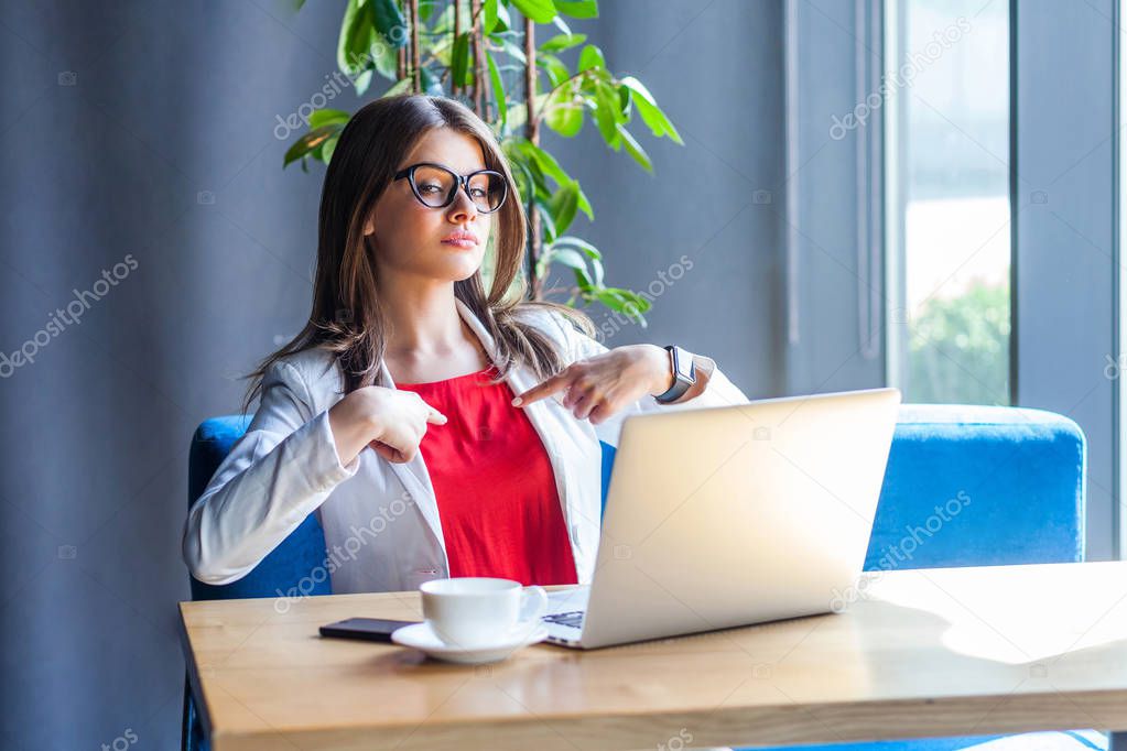 proud beautiful stylish brunette young woman in glasses looking at laptop screen on video call and pointing herself while sitting at table in cafe, freelancing concept   