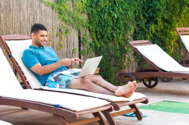 sürpriz pozitif sakallı genç yetişkin serbest adam mavi t-shirt ve şort havuz kenarında şezlong üzerinde yatan ve dizüstü bilgisayar ekranında parmak işaret 