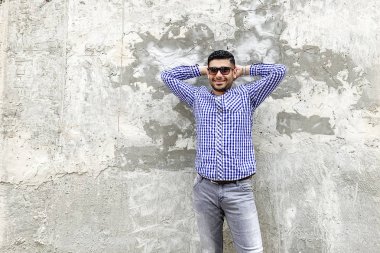 damalı mavi gömlek ve güneş gözlüğü ayakta mutlu başarılı sakallı genç adam ve beton gri duvara karşı dişli gülümseme ile kamera bakıyor