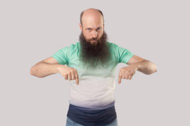 açık yeşil t-shirt uzun sakallı ciddi orta yaşlı kel adam aşağı işaret ve gri arka planda kamera bakıyor