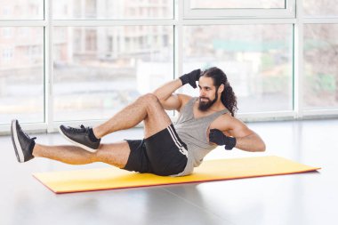 Uzun kıvırcık saçlı yakışıklı, kilolu, genç, kaslı sporcu paspasın üzerinde uzanıyor ve vücut çalışırken kollarını ve bacaklarını çaprazlayarak sallıyor.,