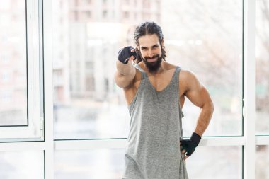 Pencereyakınında dururken kıvırcık uzun saçlı ve kameraya parmak işaret memnun genç yetişkin kas eğitmen adam 