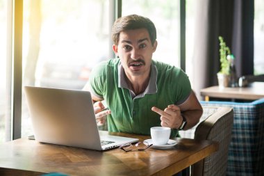 Young angry crazy businessman in green t-shirt sitting with laptop in cafe and looking at camera while showing middle finger fuck signs 