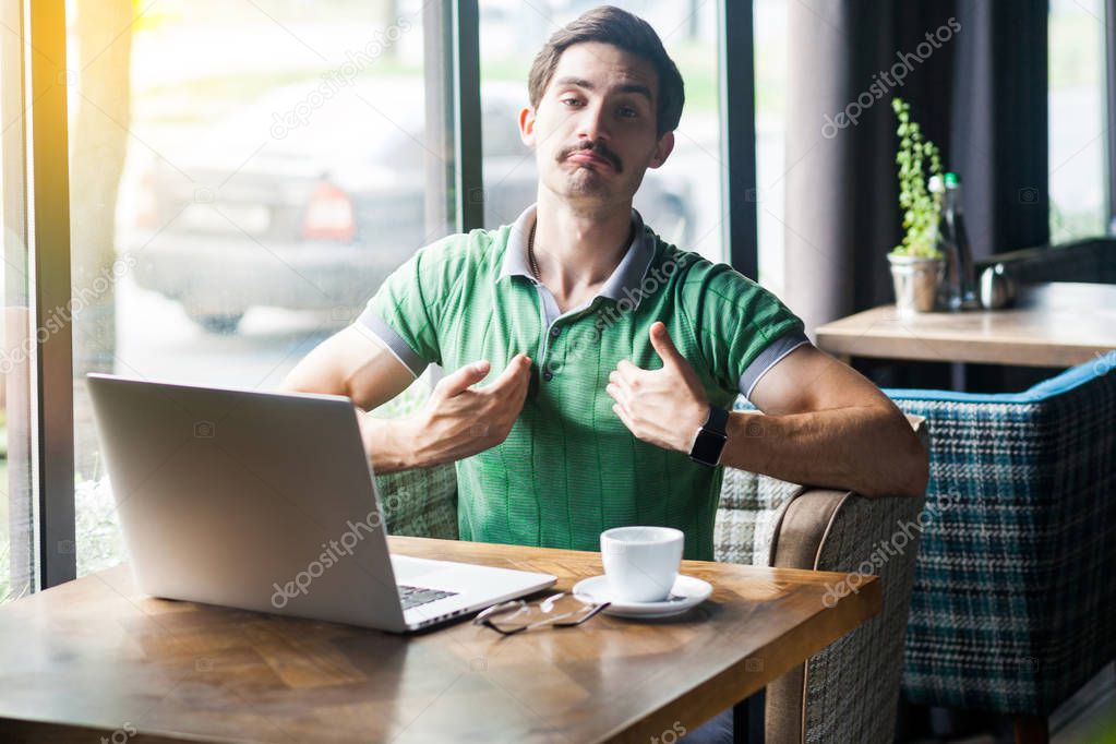 Young proud haughty businessman in green t-shirt sitting with laptop in cafe and looking at camera with proud confident face. business and freelancing concept
