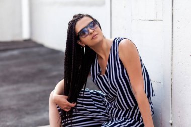 Siyah Afro-Amerikan saçı olan esmer bir kadın çizgili elbiseli, güneş gözlüklü bir kadın sokakta duvarın yanındaki duvara bakarken oturup saçlarını tutuyor. 