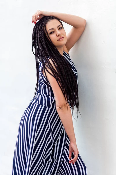 serious young brunette woman with black afro american dreadlocks hairstyle in striped dress posing and looking at camera while leaning at wall outdoor