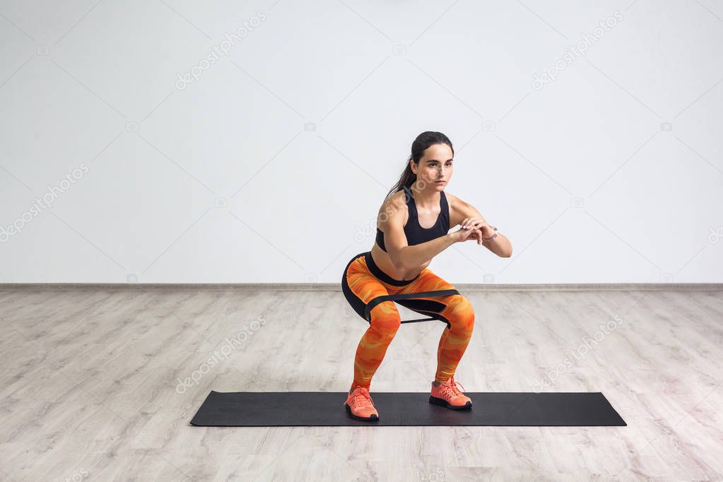 young sporty beautiful woman in black top and orange leggings doing squatting with elastic resistance band while standing on mat in gym
