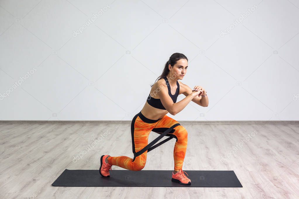 young sporty healthy attractive woman in black top and orange leggings doing lunge with elastic resistance band while standing on mat in gym