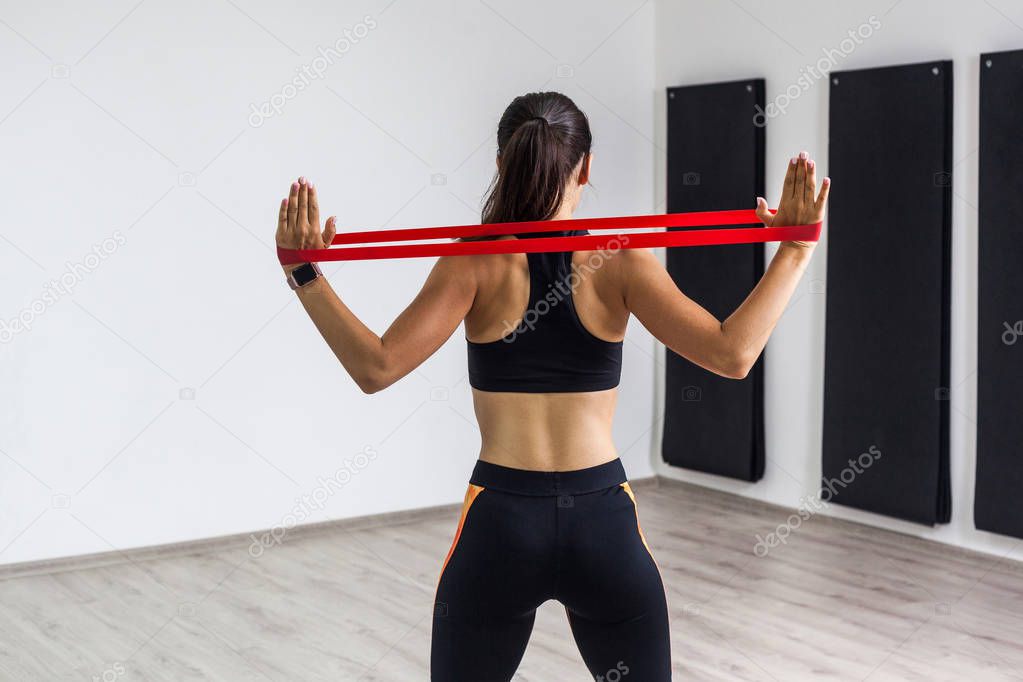 back view of athletic body fitness beautiful woman in black top and orange leggings performing exercises for muscles of back and hands with resistance band while standing in gym