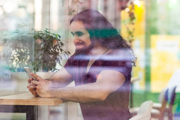 Side view through window of happy handsome athlete man with curly long hair sitting on cafe and holding smartphone while reading social media