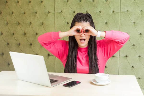 surprised young woman with black dreadlocks in pink blouse showing binoculars hands gesture on eyes and looking at camera with opened mouth while sitting at table with laptop in cafe