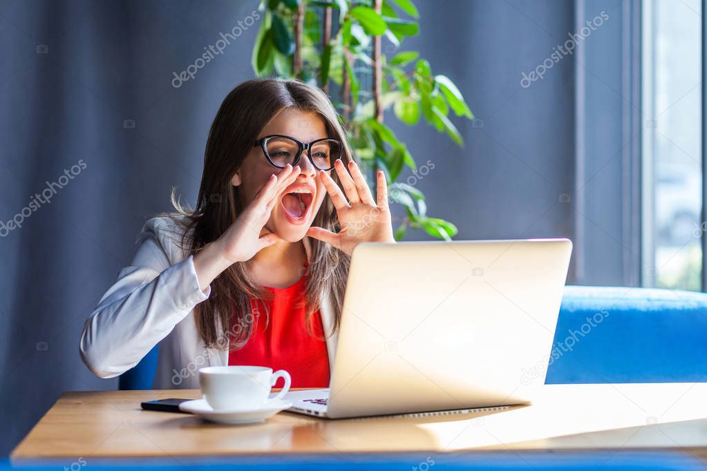 angry brunette young woman in glasses looking at laptop screen on video call and screaming while sitting at table in cafe, freelancing concept 