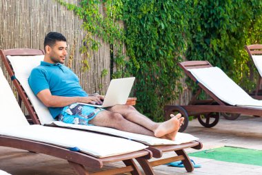zevk yakışıklı sakallı genç yetişkin serbest adam mavi t-shirt ve şort havuz kenarında dizüstü bilgisayar ile rahat daybed yalan ve online çalışma 