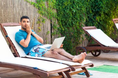 sürpriz sakallı genç yetişkin freelancer adam mavi t-shirt ve şort rahat havuz kenarında dizüstü bilgisayar ile şezlong üzerinde yatan ve büyük gözlü kamera bakıyor 