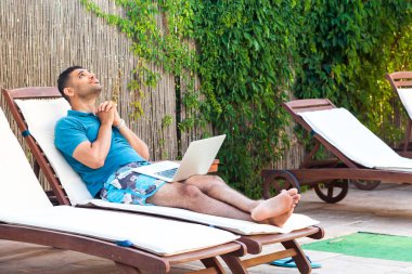 mavi t-shirt ve şort havuz kenarında dizüstü bilgisayar ile rahat şezlong üzerinde yatan yakışıklı sakallı genç yetişkin dua adam yalvaran ve ona tanrı yardım etmekten mutluluk 