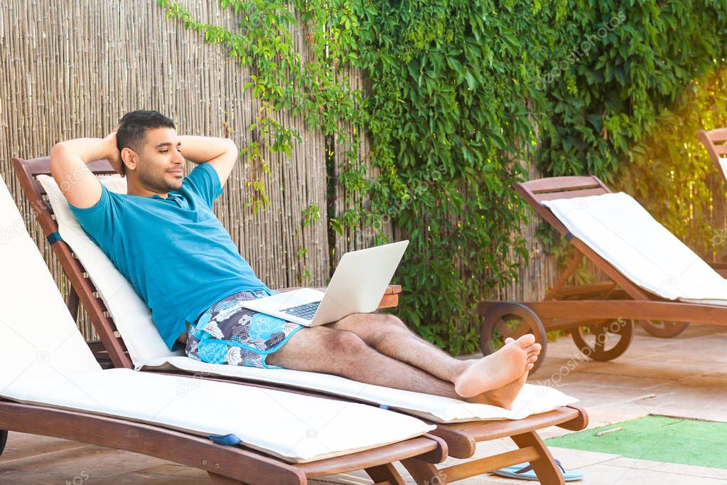 pleasure bearded young adult freelancer man in blue t-shirt and shorts lying on cozy daybed with laptop on poolside and watching film online 