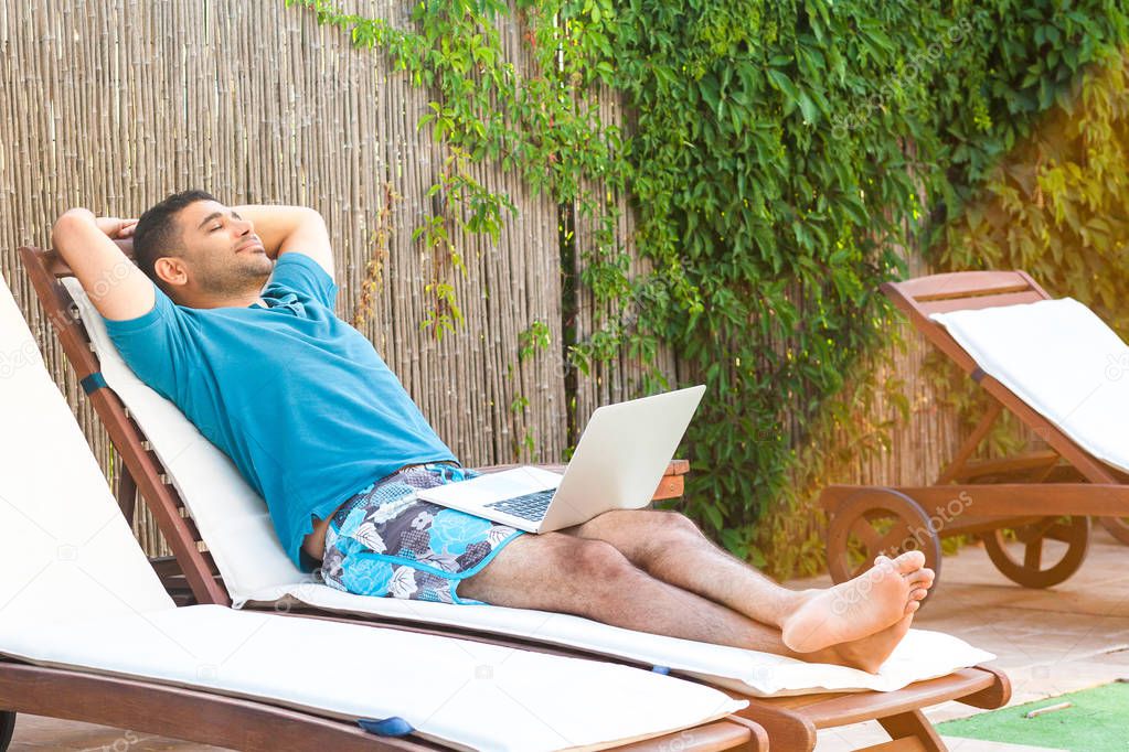 pleasure bearded young adult freelancer man in blue t-shirt and shorts lying on cozy sunbed with laptop and dreaming 