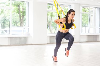 Siyah spor giysili güzel bir kadın şınav çekiyor ve spor salonunda trx fitness kayışlarıyla göğsünün üst kısmını eğitiyor.