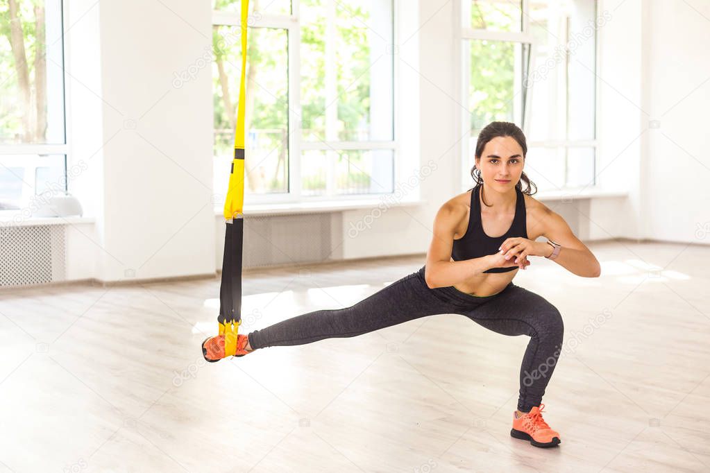 Young attractive sporty woman in black sportswear doing lunges and stretching legs while holding leg hanging on trx near window in gym 