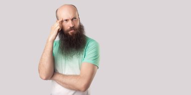 açık yeşil t-shirt ayakta uzun sakallı düşünceli orta yaşlı kel adam ayakta ve gri arka plan üzerinde uzağa bakıyor.