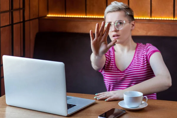 aggressive unhappy young woman freelancer with blonde short hair pleasing make pause and showing gesture stop with palm while sitting at table with laptop in cafe, freelancing concept