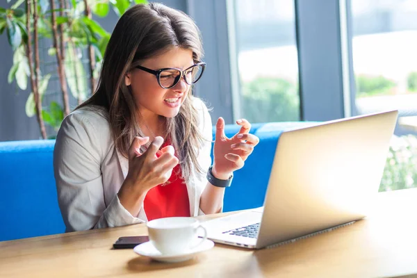 angry beautiful stylish brunette young woman in glasses looking at laptop screen with aggressive face and clenching teeth while sitting at table in cafe, freelancing concept