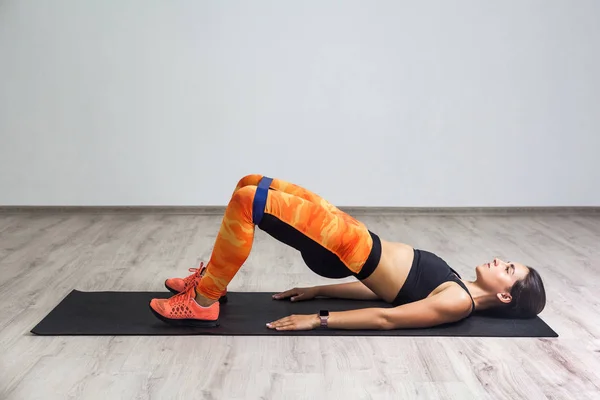 Spor kıyafetli, spor kıyafetli, güçlü beyaz bir kadının spor salonunda elastik bant kullanırken mindere uzanmış, bacak ve kalça kaslarını çalıştırması yan görünüm.