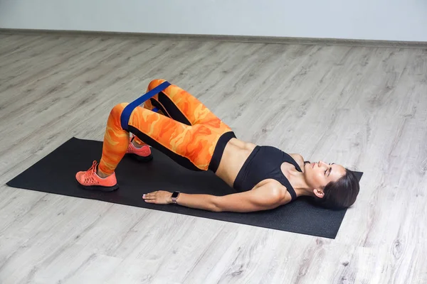 Spor kıyafetli, spor kıyafetli, güçlü beyaz bir kadının spor salonunda elastik bant kullanırken mindere uzanmış, bacak ve kalça kaslarını çalıştırması yan görünüm.